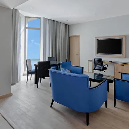 Makarem Umm Al Qura Hotel Mecca Exterior photo The photo shows a modern living area or hotel suite. It features a light-colored wooden floor and large windows allowing natural light to flood in. There are blue upholstered chairs arranged around a glass coffee table. A black office chair is positi