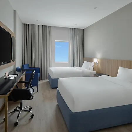 Makarem Umm Al Qura Hotel Mecca Exterior photo The photo shows a hotel room featuring two beds with white linens and blue bases. There is a modern desk and chair positioned against one wall, with a large flat-screen television mounted above it. Natural light comes in through a window covered by s