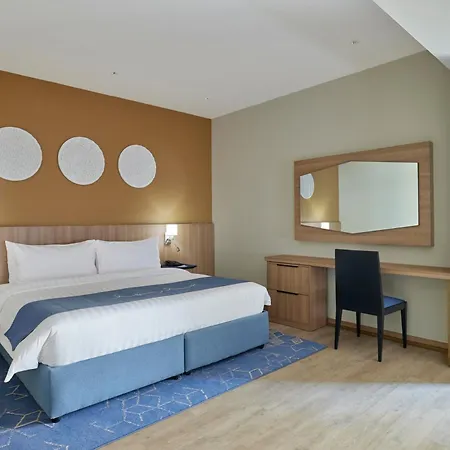 Makarem Umm Al Qura Hotel Mecca Exterior photo The photo shows a modern hotel room. It features a large bed with white linens and a blue base. On the wall above the bed are three circular decorative pieces. There is a desk with a mirror next to it, and a black chair is positioned in front of the 